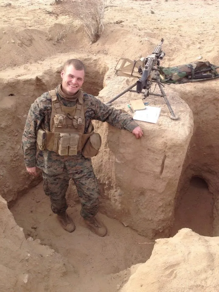 Me standing in a dug-in fighting position in the desert, with an M240B on a tripod set up beside me.